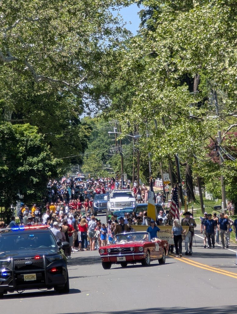 A crowd fills the road during Shrewsbury’s centennial parade, with a red vintage car and American flags leading the procession.
